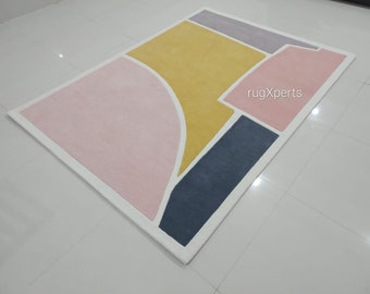 May include: Abstract geometric rug with a white border and a pink, yellow, gray, and blue color scheme. The rug has the text "rugXperts" in the upper right corner.