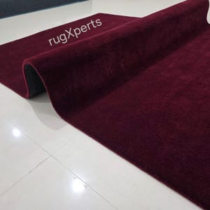 May include: A rolled-up burgundy rug with a plush texture. The rug is partially unrolled, revealing a dark gray underside. The text "rugXperts" is visible on the rug's surface. The rug is placed on a white tiled floor.