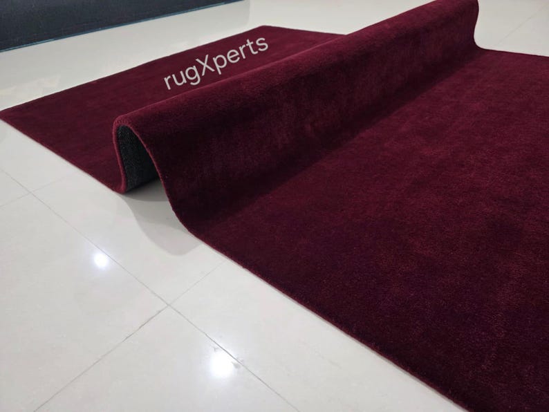 May include: A rolled-up burgundy rug with a plush texture. The rug is partially unrolled, revealing a dark gray underside. The text "rugXperts" is visible on the rug's surface. The rug is placed on a white tiled floor.