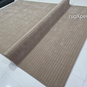 Op de afbeelding: Een beige, rechthoekig tapijt met een geribbelde textuur, gedeeltelijk opgerold om de onderkant te onthullen. Het tapijt ligt op een betegelde vloer. De tekst "rugXperts" is zichtbaar op het oppervlak van het tapijt.