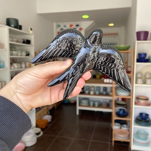 Può includere: Figurina di rondine in ceramica nera lucida con ali spiegate. L'uccello presenta dettagliate trame di piume e un design elegante. La figurina è tenuta in mano, mostrando le sue dimensioni e la sua forma.