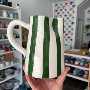 Puede incluir: Una jarra de cerámica con acabado brillante que presenta rayas verticales de color verde oscuro sobre un fondo crema. La jarra tiene un asa curva en el lado izquierdo y un borde superior ligeramente ondulado e irregular.