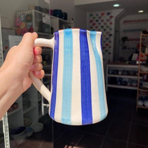 Pode incluir: Um jarro de cerâmica com um padrão de riscas verticais em tons alternados de azul escuro e azul claro sobre um fundo branco. O jarro possui uma pega curva na lateral e um rebordo largo e ligeiramente alargado com um bico vertedor.