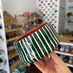 Op de afbeelding: Een keramische kom met een donkergroene buitenkant met verticale witte strepen en een gladde, terracotta-kleurige binnenkant. De kom heeft een ronde, licht taps toelopende vorm en een glanzende afwerking, die een duidelijk contrast laat zien tussen het donkere buitenpatroon en de warme, aardse tint van het binnenoppervlak.