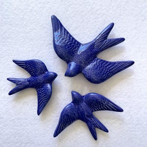 Może przedstawiać: Trzy błyszczące, ciemnoniebieskie ceramiczne ozdoby ścienne w kształcie jaskółek w locie. Każdy ptak posiada szczegółowe, formowane tekstury piór na skrzydłach i ciele, z gładkim, odblaskowym wykończeniem szkliwa. Zestaw zawiera trzy ptaki.