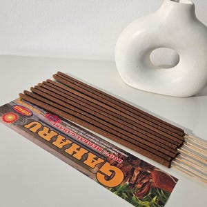 May include: A collection of brown incense sticks with wooden stems, arranged on a white surface. A package with the text "GAHARU" is visible. A white, abstract vase with a hole is in the background.