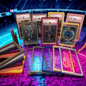 May include: A collection of basketball trading cards in protective cases, displayed on stands. The cases have various colored frames, including yellow, pink, and blue. The background features a basketball court with neon lighting.