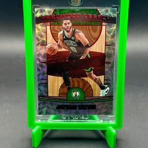 May include: A basketball trading card of Jason Tatum in a Boston Celtics uniform, displayed in a green plastic case. The card has a holographic background with the text "HARDWOOD LEGACY". The player is shown dribbling a basketball.