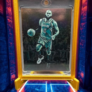 May include: A basketball trading card featuring LeBron James in a teal Lakers jersey, dribbling a basketball. The card is encased in a gold-coloured holder with a clear front, displayed against a blue and red background.