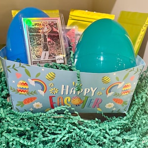 Può includere: Un cestino pasquale con un uovo blu e verde acqua, una carta da basket e un riempitivo di carta verde. Il cestino è decorato con le parole "Buona Pasqua", arcobaleni e uova di Pasqua.