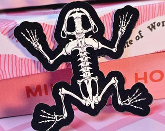 Op de afbeelding: Een zwarte sticker in de vorm van een kikker skelet met witte bot details. De sticker is geposeerd met zijn armen en benen uitgestrekt. De achtergrond bevat roze en wit gestreepte dozen met de woorden "MI" en "HONEY".