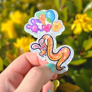May include: A colorful sticker featuring a cartoon snake with bunny ears, holding balloons. The snake is orange with a black outline, and the balloons are various colors. The sticker has a glittery background.