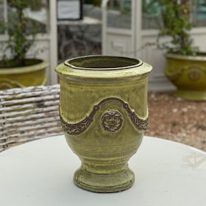 May include: A handcrafted, light green ceramic planter with a decorative brown floral design. The planter has a wide opening and a pedestal base. The background features a greenhouse and other planters, suggesting a garden or outdoor setting.