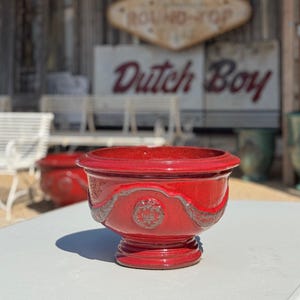 May include: A vibrant red ceramic planter with a decorative embossed design. The planter has a wide, shallow bowl shape and sits on a pedestal base. The background includes a sign that says "Dutch Boy".
