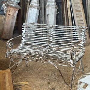 May include: An antique white metal bench with a slatted seat and backrest. The bench has curved armrests with decorative scrollwork. The paint is distressed, revealing the metal underneath. The bench is outdoors, with architectural salvage in the background.