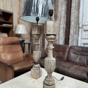 May include: Two antique wooden table lamps with a distressed finish. Each lamp features a square base, a turned wooden column, and a black lampshade. The lamps are placed on a light-colored table, with a leather sofa in the background.