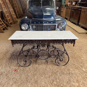 May include: An antique marble-topped table with ornate, dark metal scrollwork legs. The rectangular tabletop is white, and the metal base features intricate designs. A vintage blue Volvo truck is in the background.