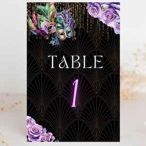 Peut inclure: Carte de numéro de table noire et dorée avec un masque de mascarade violet et vert, des fleurs violettes et le texte "TABLE 1" en blanc.