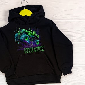 May include: Black hooded sweatshirt featuring a green monster truck graphic and the name "HUDSON" printed on the front.