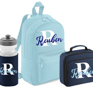 May include: A blue backpack, a blue lunch bag, and a water bottle with a blue and white insulated sleeve. All three items have the name "Reuben" written on them in white letters with a blue outline.