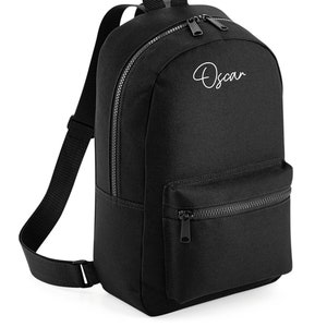 Pode incluir: Mochila de lona preta com um bolso frontal com zíper e um nome bordado em branco "Oscar".