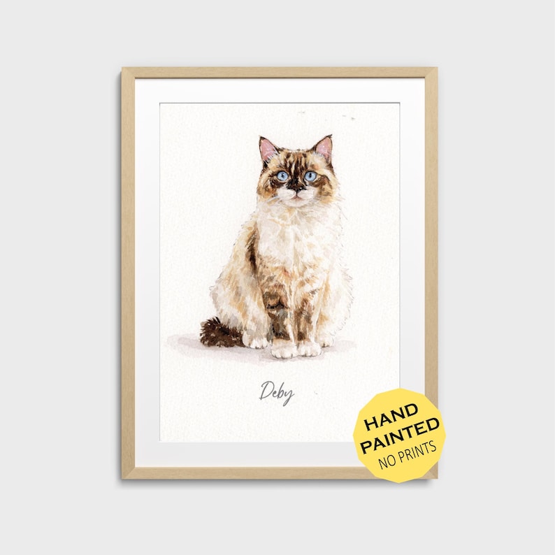 May include: Watercolor painting of a Ragdoll cat with blue eyes, sitting upright. The cat has brown and white fur. The artwork is in a light wooden frame. A yellow sticker in the bottom right corner reads "HAND PAINTED NO PRINTS". The cat's name, "Deby", is written below.