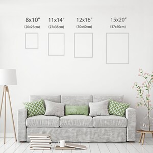 May include: Four white picture frames of different sizes hanging on a white wall. The frames are labeled with their dimensions in inches and centimeters. The frames are hanging above a gray couch with green throw pillows.