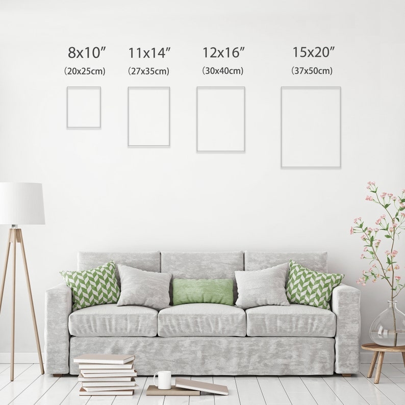 May include: Four white picture frames of different sizes hanging on a white wall. The frames are labeled with their dimensions in inches and centimeters. The frames are hanging above a gray couch with green throw pillows.
