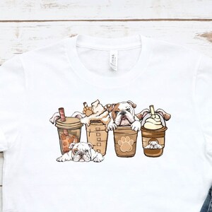 May include: White t-shirt with a graphic of four bulldogs wearing coffee cups. The bulldogs are wearing different types of coffee drinks, including a latte, a cappuccino, and a cold brew. The t-shirt is laid out on a wooden surface with a pair of white sneakers and a pair of blue jeans.