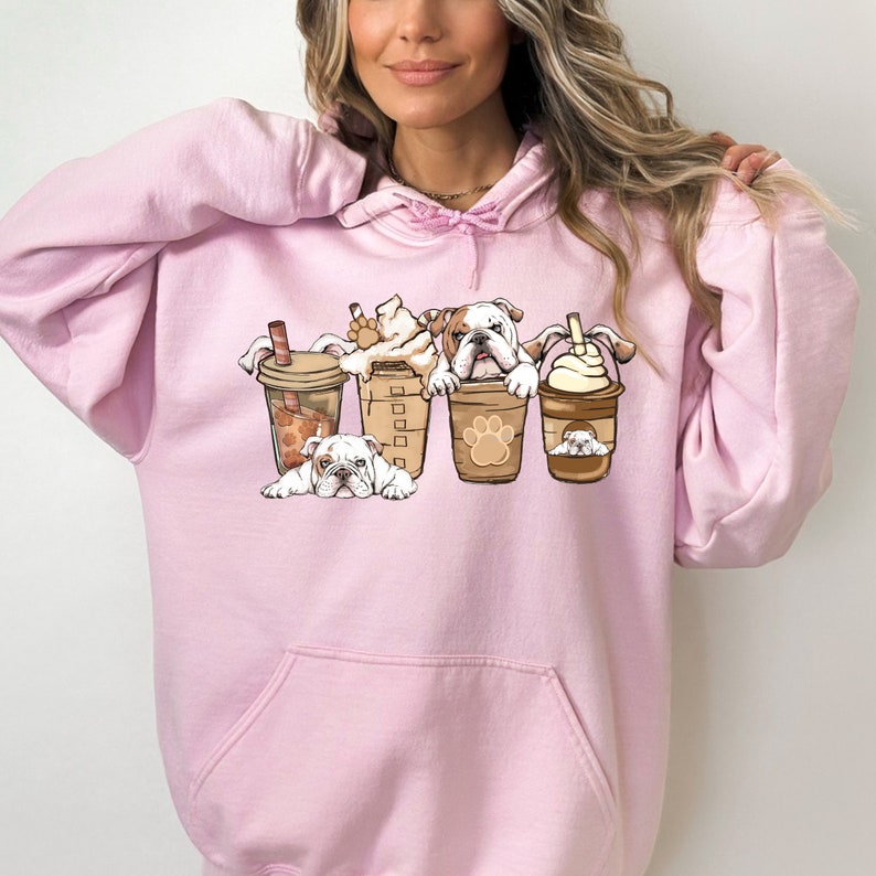 May include: Pink hoodie with a graphic of four bulldogs in coffee cups. The bulldogs are wearing brown collars and have white fur. The coffee cups are filled with different types of coffee drinks.
