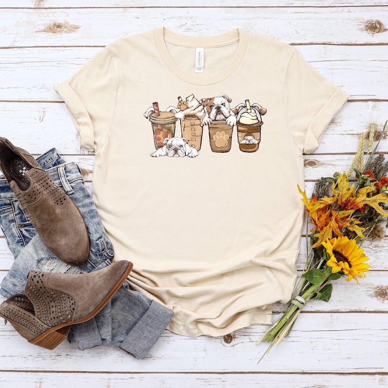 May include: A beige t-shirt with a graphic of four bulldogs peeking out of coffee cups. The bulldogs are white with brown spots and are wearing a variety of coffee cup accessories.