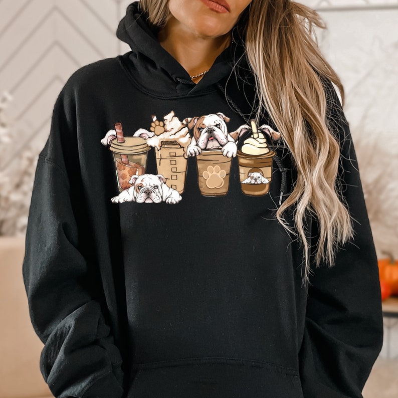 May include: Black hoodie with a graphic of four bulldogs in coffee cups. The bulldogs are white with brown spots and are looking at the viewer. The coffee cups are brown with white lids and have a paw print design.