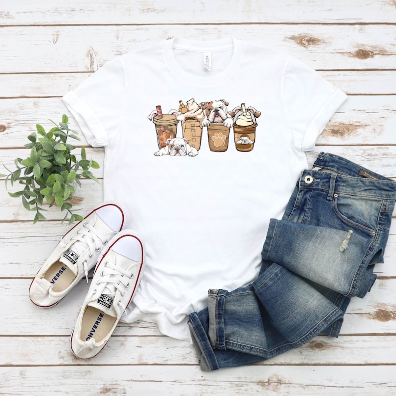 May include: White t-shirt with a graphic of four bulldogs wearing coffee cups. The bulldogs are wearing different types of coffee drinks, including a latte, a cappuccino, and a cold brew. The t-shirt is laid out on a wooden surface with a pair of white sneakers and a pair of blue jeans.