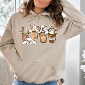 May include: A light beige hoodie with a graphic of four bulldogs peeking out of coffee cups. The bulldogs are white with brown spots and have black noses. The coffee cups are brown with white lids and have a paw print design.