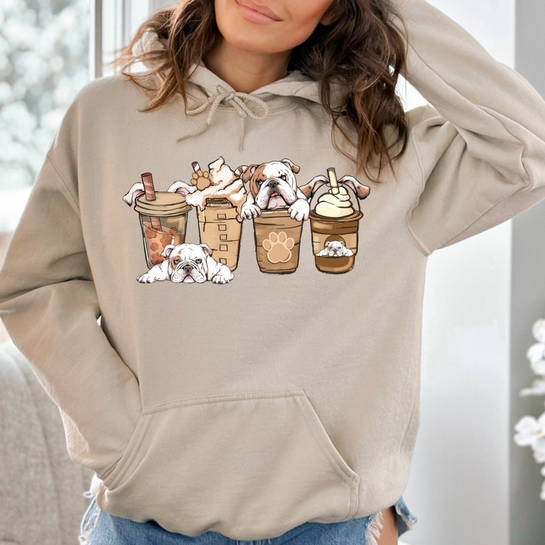 May include: A light beige hoodie with a graphic of four bulldogs peeking out of coffee cups. The bulldogs are white with brown spots and have black noses. The coffee cups are brown with white lids and have a paw print design.