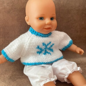 May include: A baby doll wearing a white knitted sweater with a blue snowflake design and white pants.
