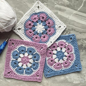 May include: Three crocheted squares in shades of pink, blue, and white. The squares have a floral design in the center and are made with a white crochet hook.