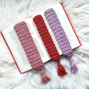 May include: Three handmade crochet bookmarks in shades of pink, red, and lavender, each with a matching tassel. The bookmarks are placed in an open book with red covers, resting on a white, fluffy surface.