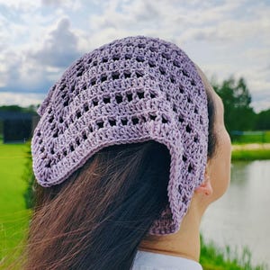 May include: A lavender crochet head covering with an open weave design. The head covering is worn on the head and covers the top of the head and the back of the head. The background includes a lake, green grass, and a cloudy sky.