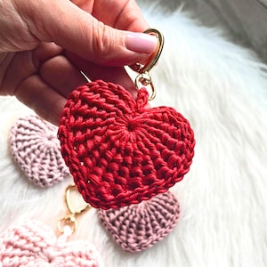 May include: A red crocheted heart keychain with a gold-colored clasp is held up, with three other crocheted hearts in shades of pink in the background. The heart-shaped keychains are handmade and have a textured appearance.