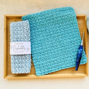 May include: Two handmade crocheted dishcloths in blue and light blue colors, with a label that reads "Handmade Dishcloths 100% cotton yarn", displayed on a wooden tray with a crochet hook.