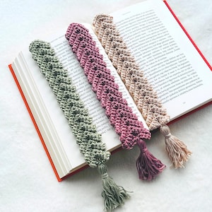 May include: Three crocheted bookmarks with tassels in green, pink and beige. The bookmarks are made with a textured stitch and have a scalloped edge.