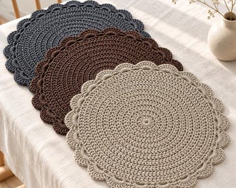 May include: Three circular crochet placemats arranged in a stack, featuring a scalloped edge design. The placemats are coloured in dark grey, dark brown, and beige, each crafted with a dense, textured stitch pattern in a round formation.