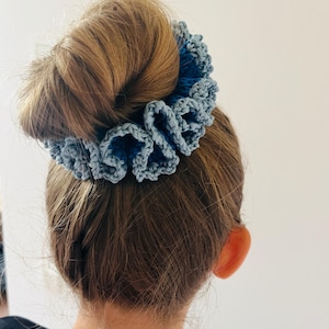 Puede incluir: Una scrunchie de ganchillo azul claro, con un diseño con volantes, se usa en un moño. La scrunchie está hecha de un hilo suave y texturizado. El cabello es de color marrón claro.
