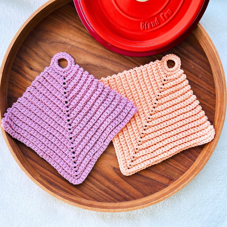 Op de afbeelding: Twee handgemaakte gehaakte pannenlappen op een houten dienblad. E&eacute;n is lavendelkleurig, de andere perzik. Beide zijn vierkant met een lus om op te hangen. Een rood ge&euml;mailleerd gietijzeren pannendeksel staat op de achtergrond.