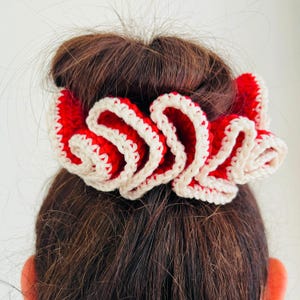 May include: A red and white crocheted scrunchie, with a ruffled design, is shown in a close-up shot. The scrunchie is made of red yarn with white trim. The scrunchie is worn in a bun. The hair is dark brown.