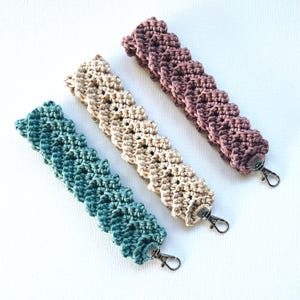 Puede incluir: Tres pulseras de ganchillo hechas a mano en verde azulado, beige y malva. Cada pulsera presenta un diseño texturizado y festoneado y un cierre plateado. Las pulseras se muestran sobre un fondo blanco.