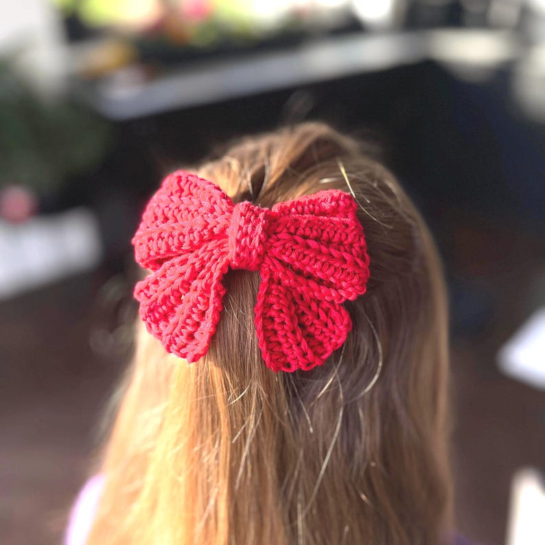 May include: A bright red crochet hair bow featuring a textured, ribbed stitch pattern. The bow is constructed with a central knot detail and two symmetrical loops on each side, creating a full, dimensional shape secured in long hair.