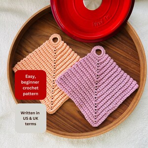 Op de afbeelding: Twee handgemaakte gehaakte pannenlappen in perzik en lavendel, met een rood pannendeksel op de achtergrond. De pannenlappen zijn vierkant met een lus om op te hangen. Tekst op de afbeelding luidt "Easy, beginner crochet pattern" en "Written in US & UK terms."