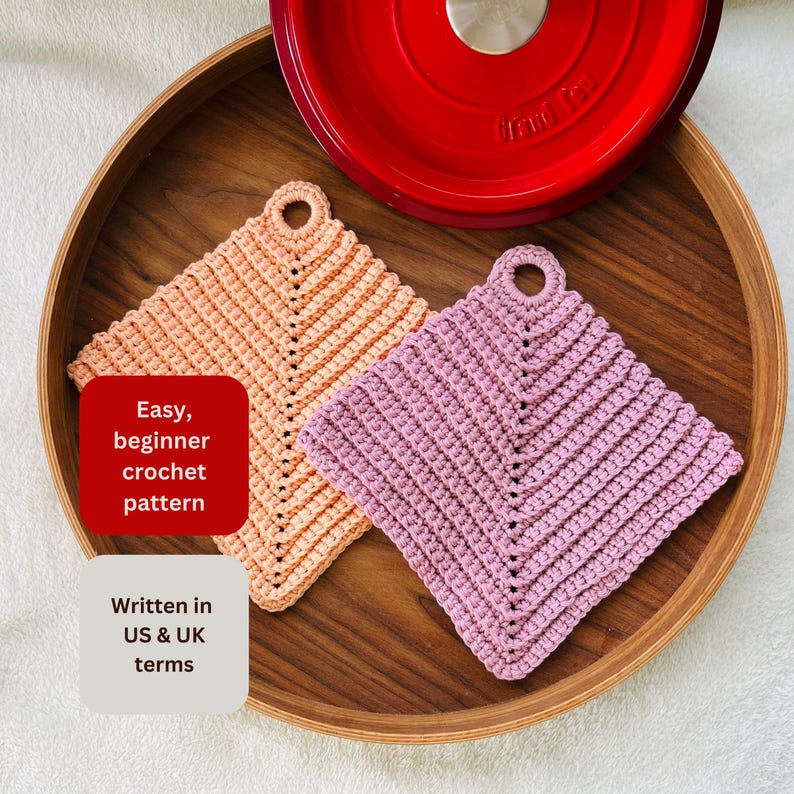Op de afbeelding: Twee handgemaakte gehaakte pannenlappen in perzik en lavendel, met een rood pannendeksel op de achtergrond. De pannenlappen zijn vierkant met een lus om op te hangen. Tekst op de afbeelding luidt "Easy, beginner crochet pattern" en "Written in US & UK terms."